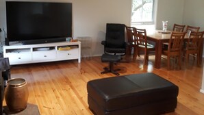 A spacious living area with a flat-screen TV, a black office chair, a dining table with wooden chairs, and a red sofa.