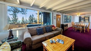 A living room with a large window, a sofa, a coffee table, and a dining area with chairs. - Rustic 6 Bedroom Cabin - Lakefront, Central in Town Location! (South Lake Tahoe)