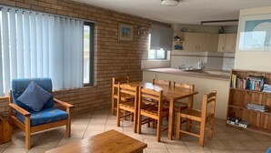 A cozy living room with a blue sofa, wooden dining table, and chairs. A kitchen area with a sink and cabinets is visible in the background. - Waikiki Unit 4 Kings Beach QLD (KINGS BEACH)