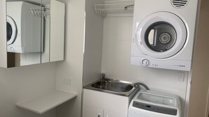 A compact, modern laundry room with a built-in washer and dryer, a stainless steel sink, and a mirror above the dryer. - Waikiki Unit 4 Kings Beach QLD (KINGS BEACH)