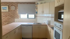 A compact kitchen with a dishwasher, a sink, a microwave, and a stove. There is a window above the sink and a clock on the wall. - Waikiki Unit 4 Kings Beach QLD (KINGS BEACH)