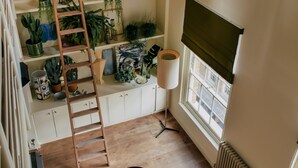Una habitación con una escalera de madera que lleva a un estante para plantas en un entrepiso, una mesa redonda para comer y una ventana con persiana enrollable.