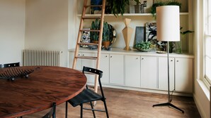 Un comedor con mesa y sillas de madera, un armario blanco con estantes, y una escalera con plantas colgantes.