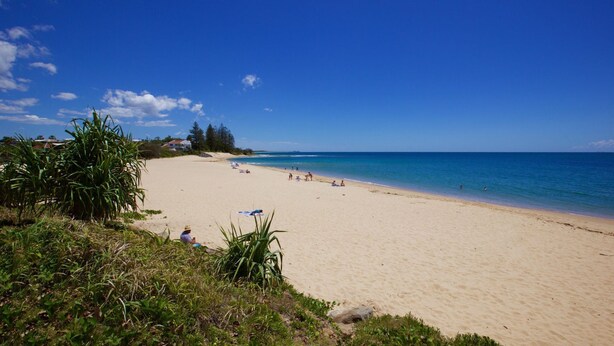 A wide sandy beach with clear blue water, a few people sunbathing, and some greenery on the left.