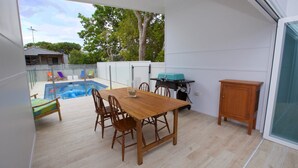 A patio with a wooden table and chairs, a barbecue grill, and a swimming pool in the background.