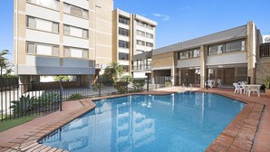 Una piscina con borde de ladrillo, rodeada por una valla y un patio con mobiliario exterior.