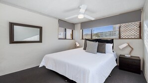 Habitación de hotel con cama, mesita de noche, lámpara de escritorio, ventilador de techo y ventanas que dan a edificios.
