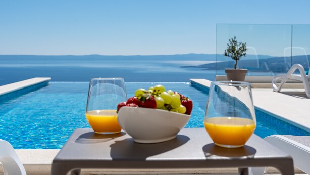 Une table placée au bord de la piscine, avec un saladier rempli de fruits et deux verres de jus d’orange, d’où l’on peut admirer une mer bleue et limpide ainsi que des montagnes lointaines.
