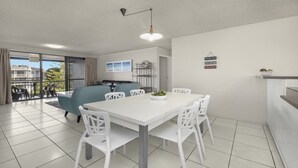 A modern living room with a white dining table and chairs, a sofa, and a balcony with outdoor seating. - Sealanes Unit 8 Kings Beach QLD (KINGS BEACH)