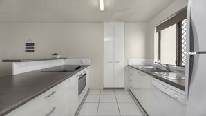A modern kitchen with white cabinets, a stainless steel sink, and a dishwasher. There is a window with a metal grid and a sign on the wall. - Sealanes Unit 8 Kings Beach QLD (KINGS BEACH)