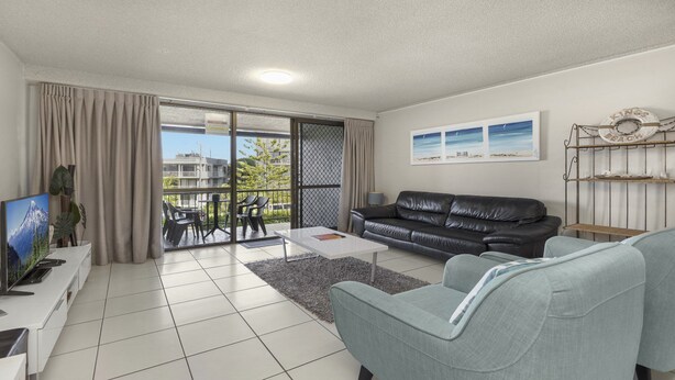 A modern living room with a black leather sofa, a grey armchair, a flat-screen TV, and a balcony with a view of buildings and greenery. - Sealanes Unit 8 Kings Beach QLD (KINGS BEACH)