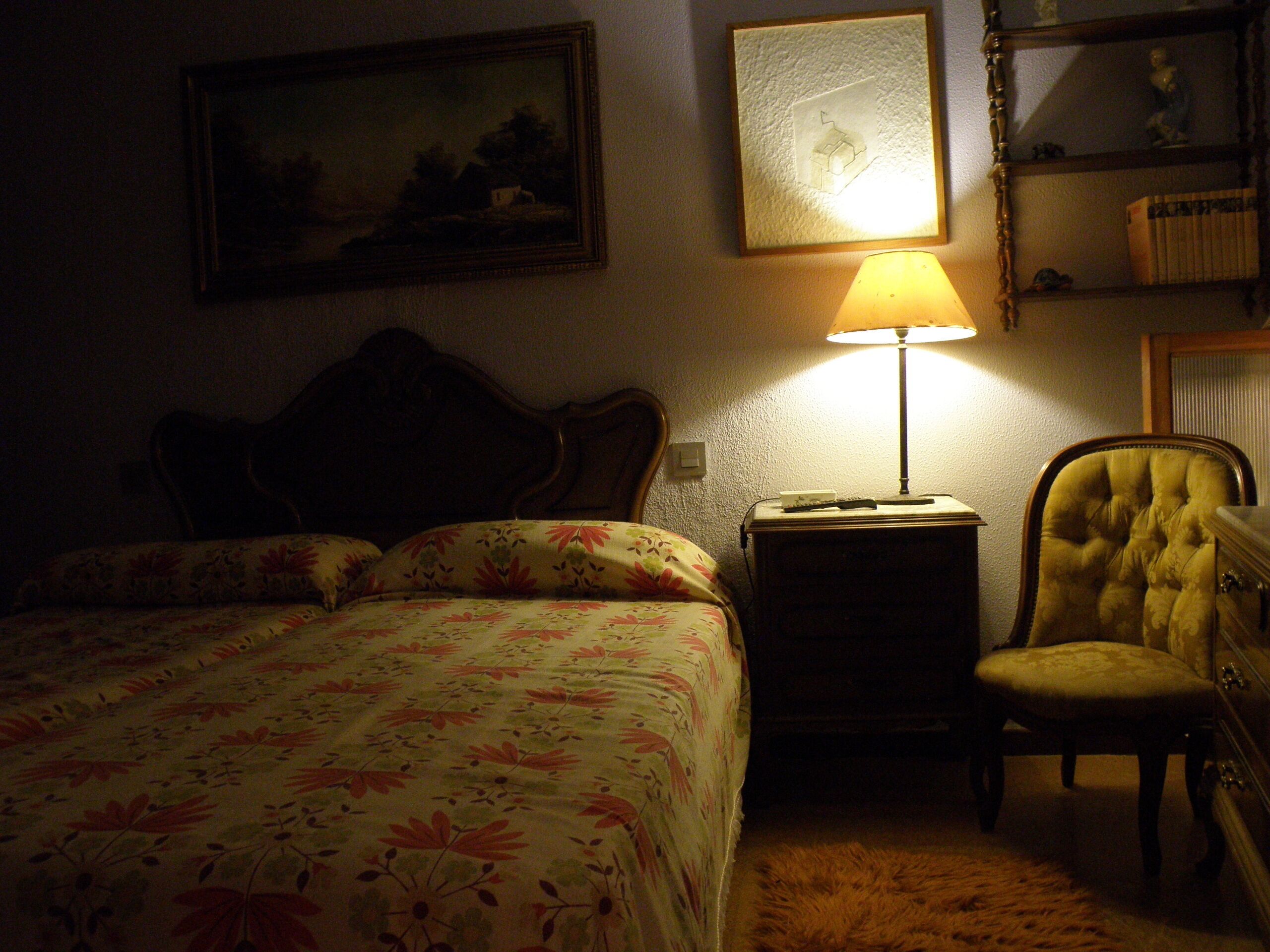 A dimly lit hotel room with a bed, a chair, a nightstand, and a lamp.