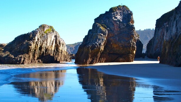 Uma praia com grandes formações rochosas e águas calmas refletindo a paisagem.