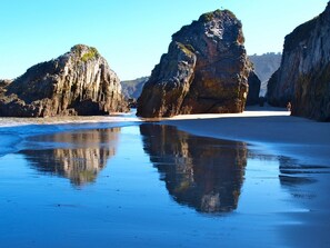 Una playa con grandes formaciones rocosas y agua tranquila que refleja el paisaje.