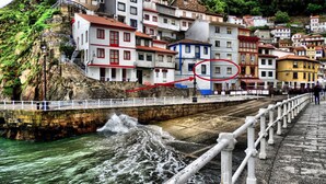 Uma vila costeira com edifícios à beira-mar, uma ponte e um penhasco rochoso.