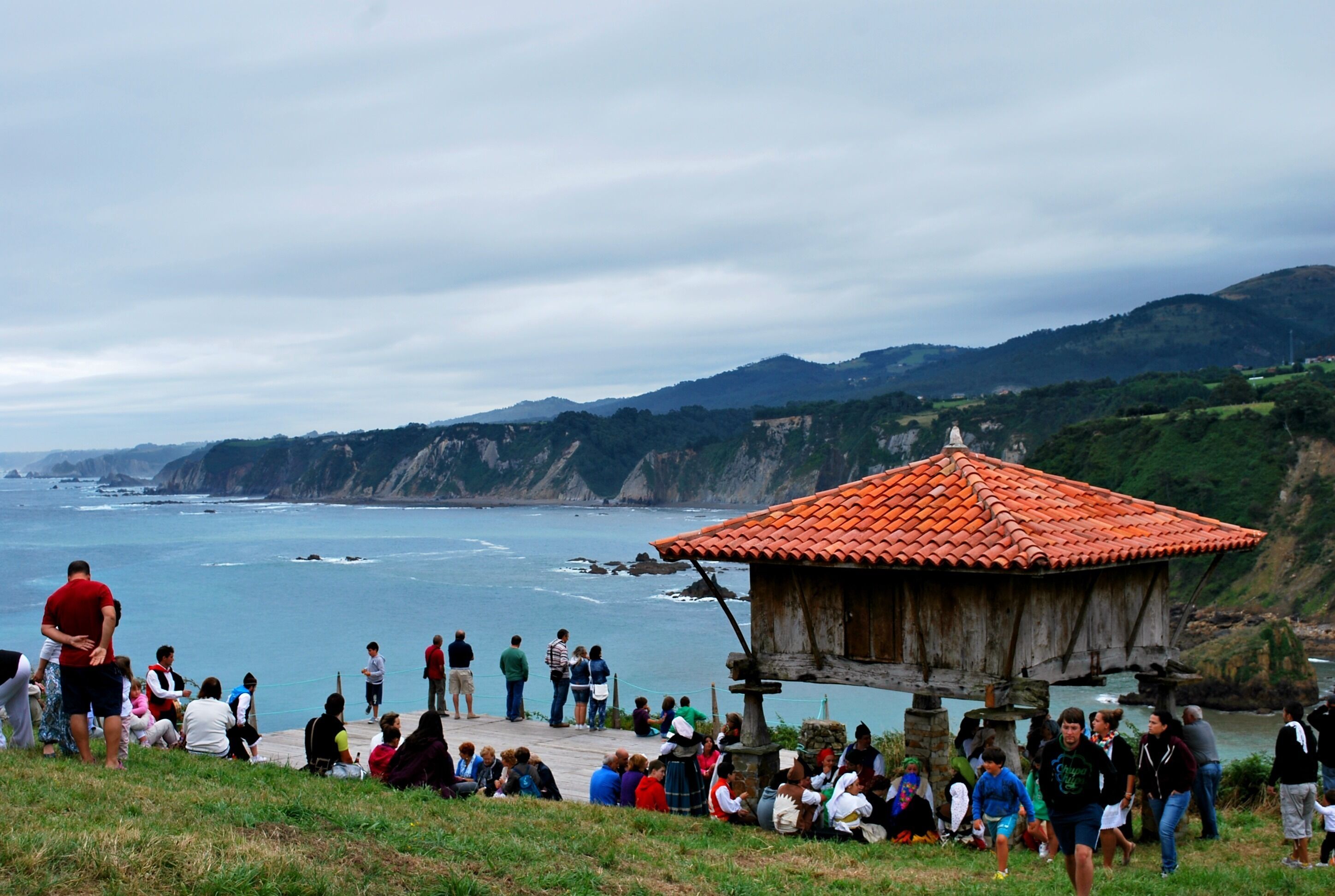 A coastal area with a wooden structure, people gathered around, and a view of the sea and hills.