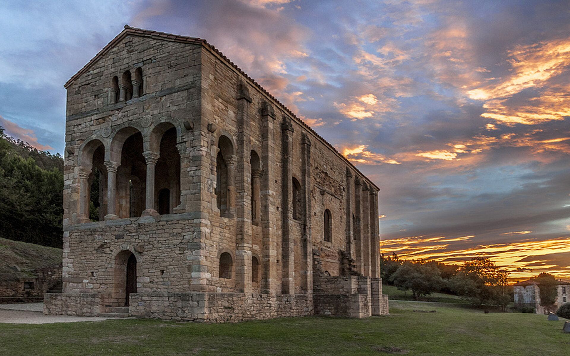 A stone building with multiple arches and a flat roof, surrounded by grass and trees, with a sunset in the background.