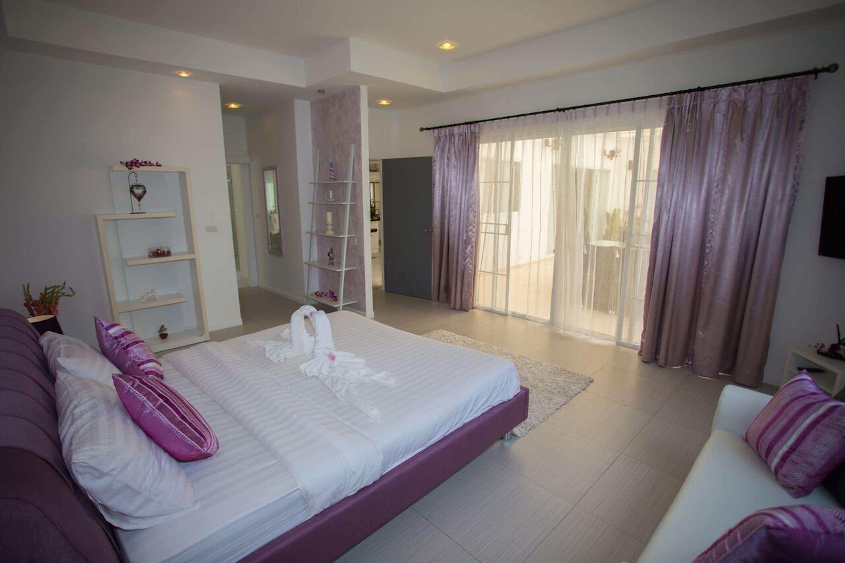 A modern bedroom with a large bed, a sofa, a TV, and a balcony with curtains.