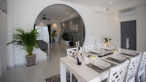 Un espace repas moderne avec une table blanche dressée pour six personnes, entourée de chaises blanches. Un pot de plante se trouve à gauche, et un mur gris avec des étagères est visible au fond.
