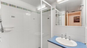 Ett badrum med en vit kakelbeklädd vägg, en duschkabin i glas, ett handfat med en blå bänkskiva och en spegel som reflekterar duschutrymmet.