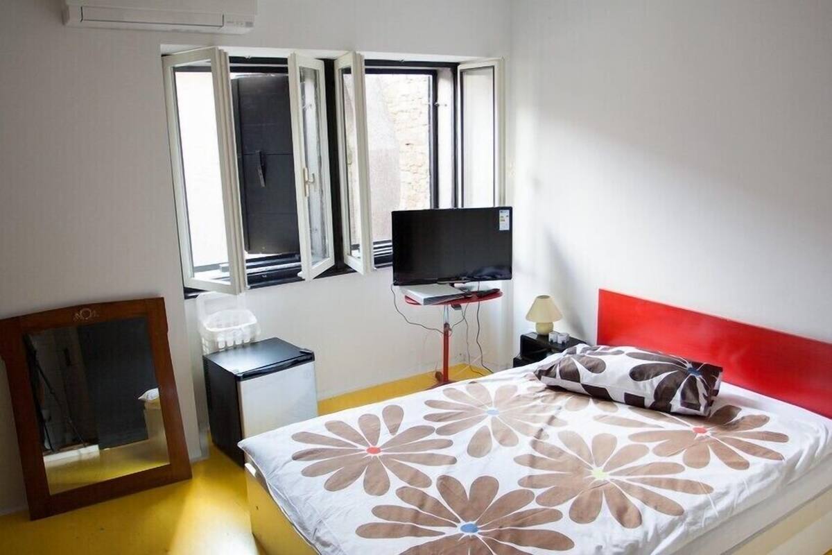 A bedroom with a bed, a TV, a desk, and a window.