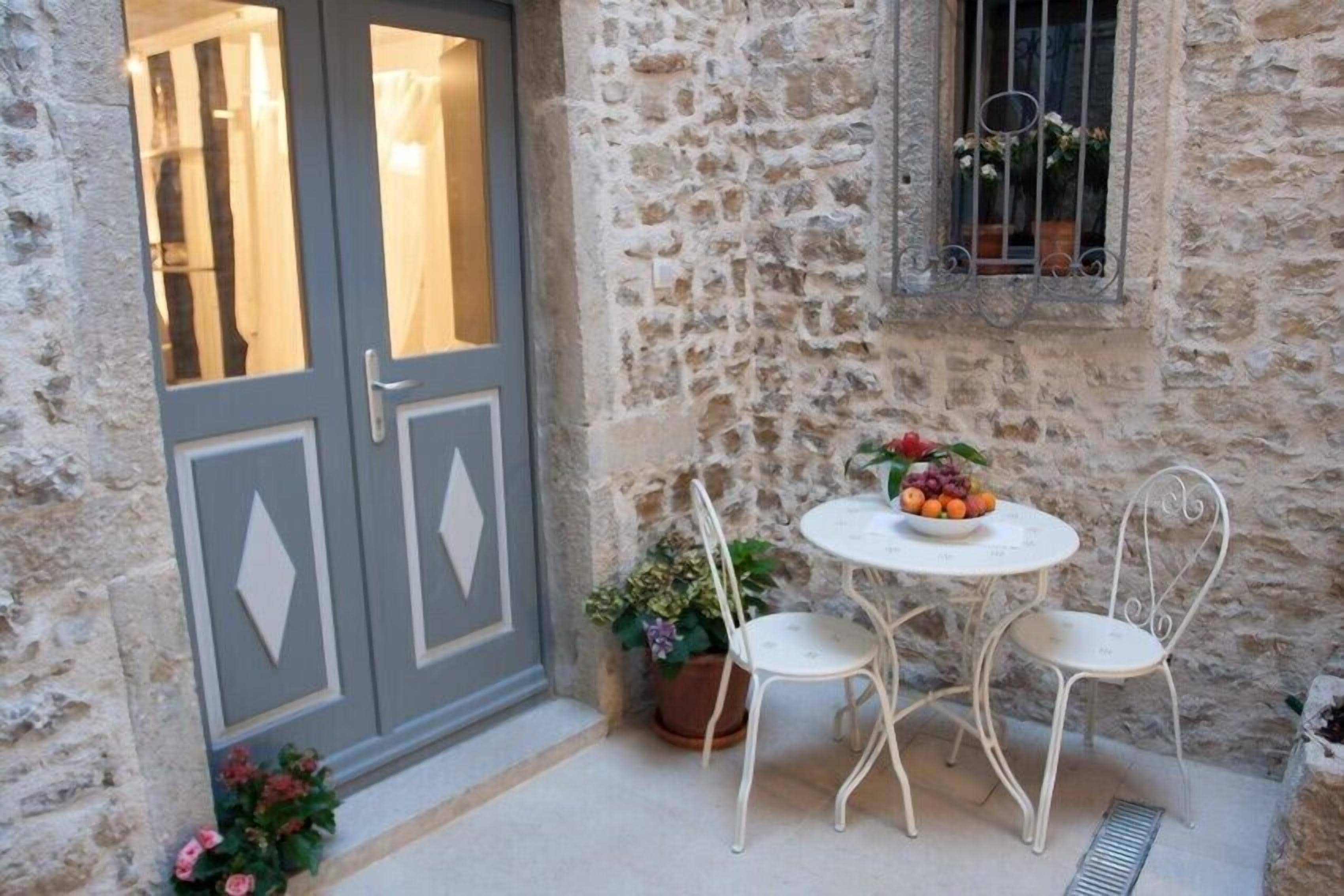 Un muro de piedra con una ventana, una puerta y una pequeña zona exterior con una mesa y dos sillas.