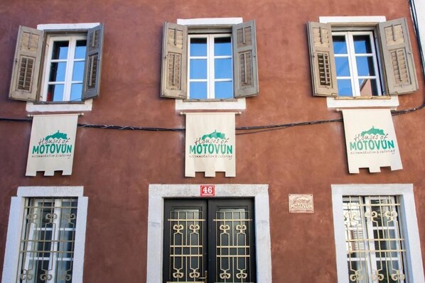 Um edifício com três janelas, cada uma com persianas de madeira. Três faixas pendem do edifício, cada uma com o texto
