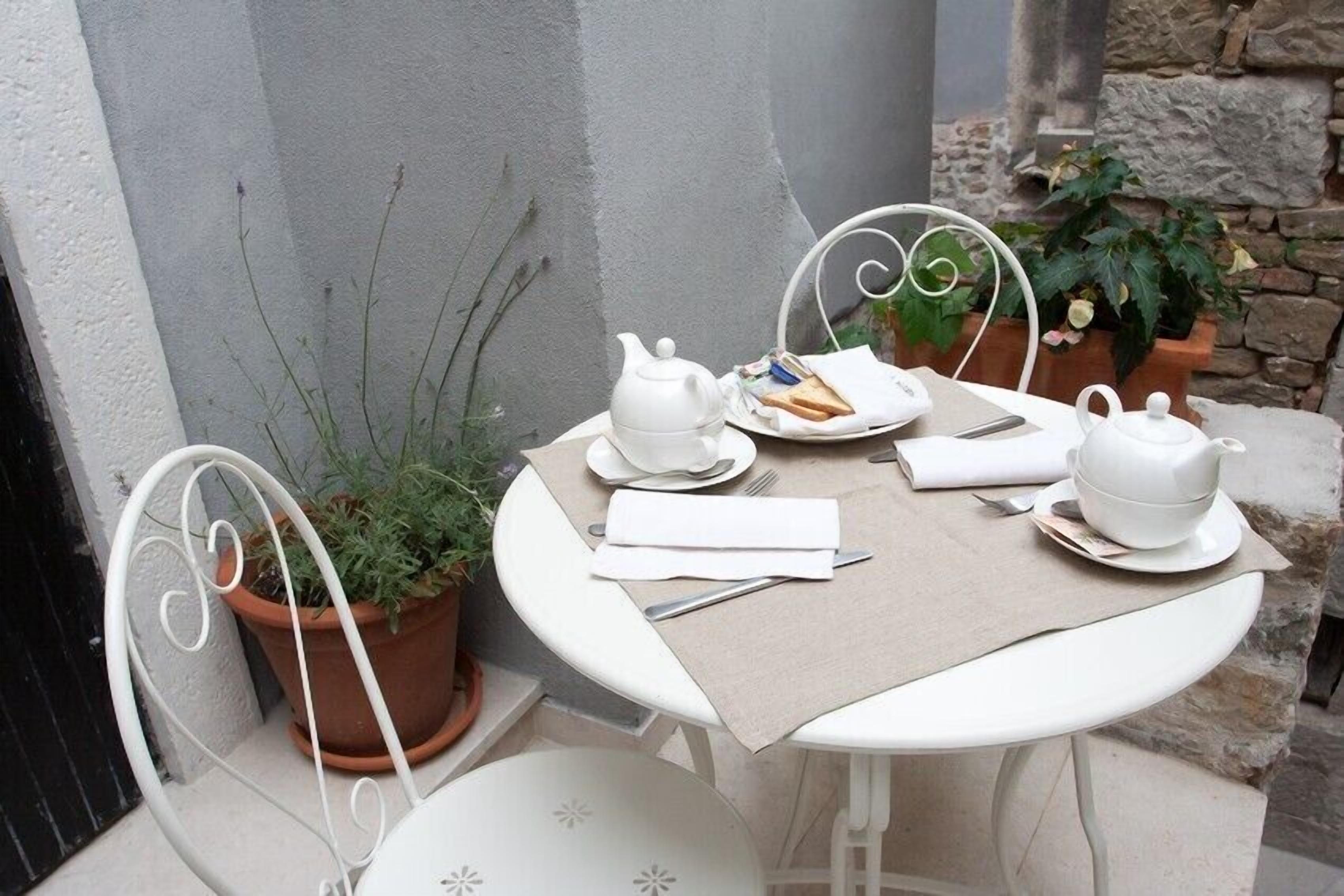 A small outdoor table set with white teapots, plates, and a napkin, surrounded by white chairs and potted plants.