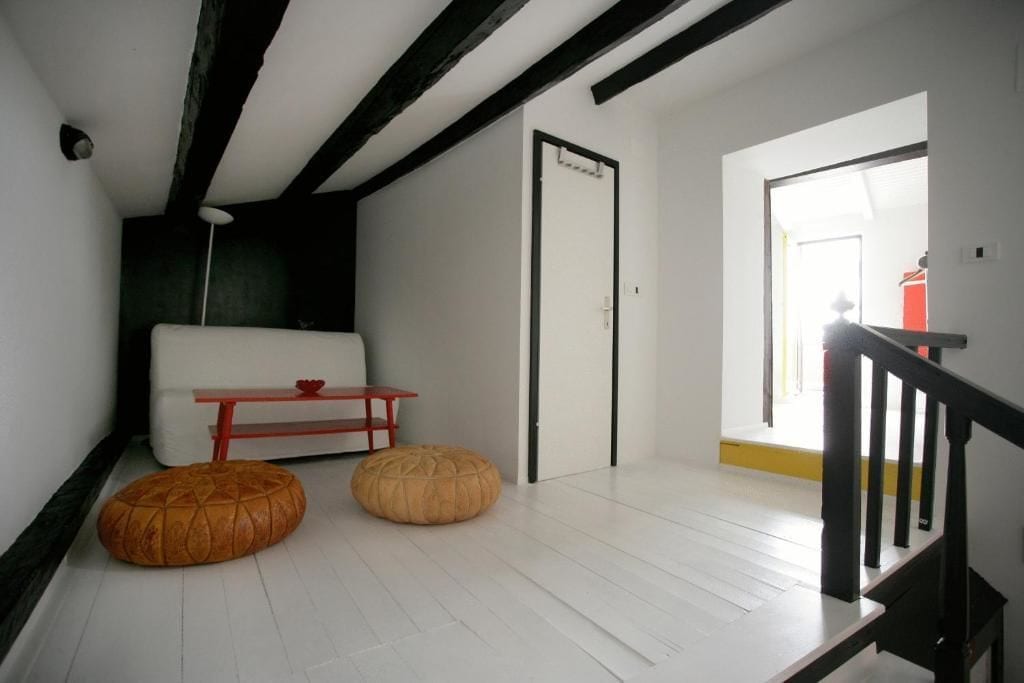 A modern room with white walls, a wooden ceiling, a white sofa, two ottomans, a red table, and a door leading to another room.