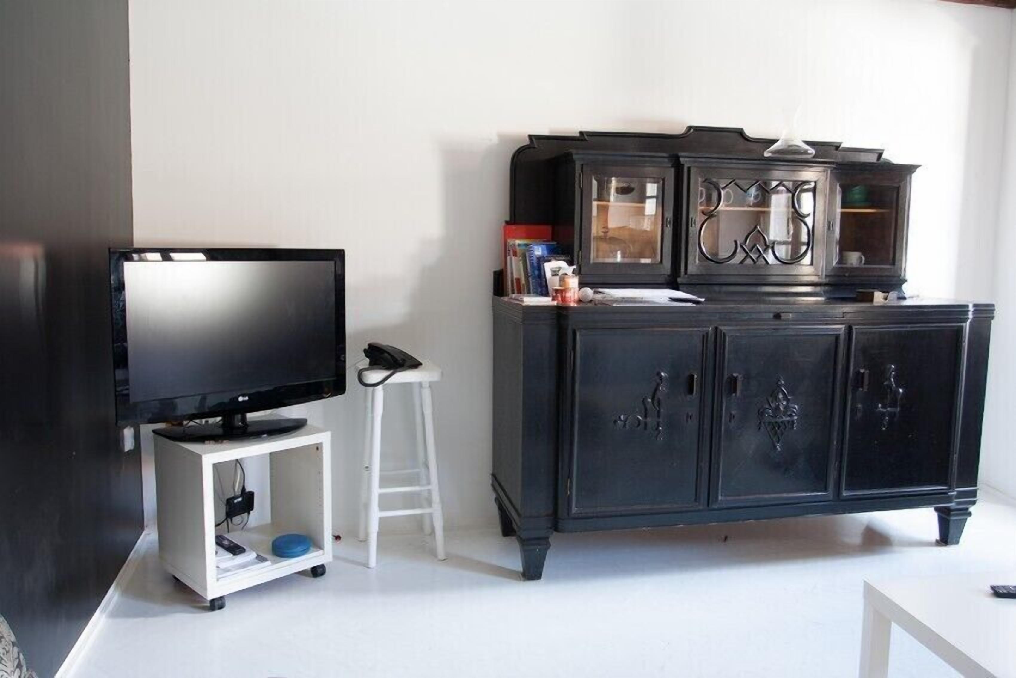A living room with a flat-screen TV on a white stand, a white stool, and a dark wooden cabinet with glass doors.