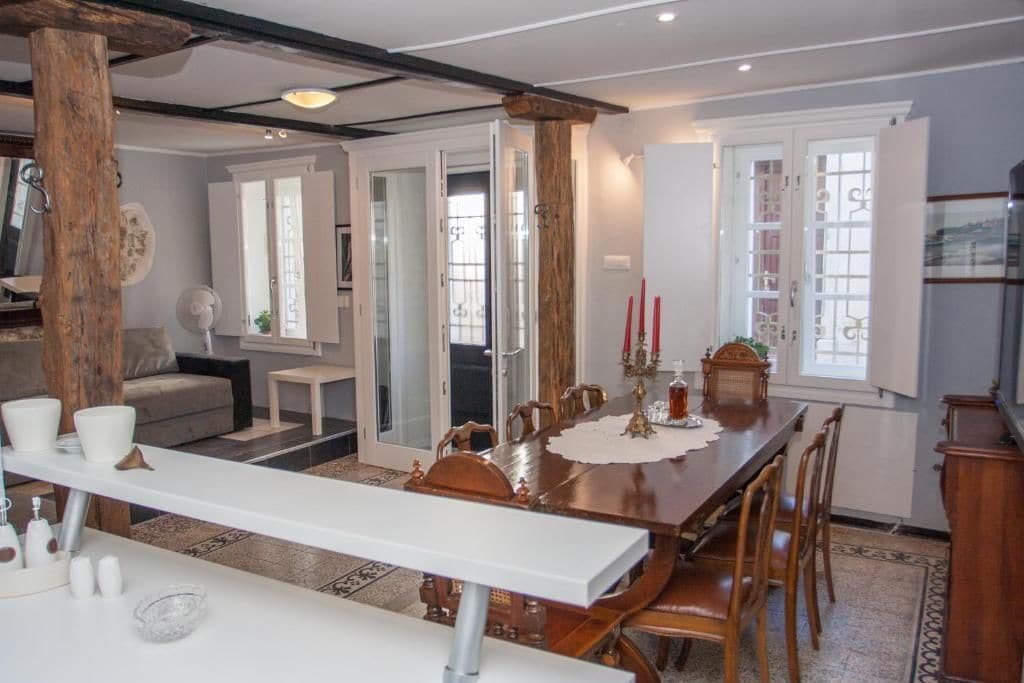 A dining area with a long wooden table, white chairs, and a white bench. There are candles on the table, and a white fan is visible in the background.