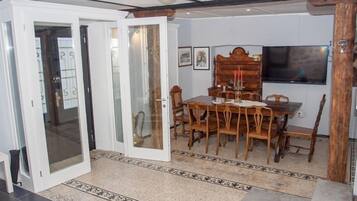 A dining area with a wooden table and chairs, a television mounted on the wall, and a patterned rug on the floor.