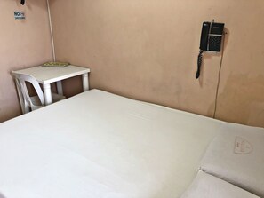 Habitación con cama individual, colchón blanco, una mesita pequeña, una silla, un cartel de no fumar y un teléfono fijo en la pared.