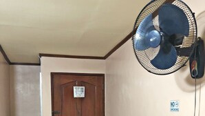 A room with a wooden door, a television on a shelf, a wall-mounted fan, and a 'No Smoking' sign.