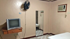 A hotel room with a television on a wooden stand, a wall-mounted fan, a bed with a white mattress, a bedside table, and a mirror.