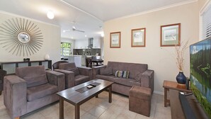 A living room with a sofa set, a coffee table, and a TV. There are framed pictures on the wall and a large mirror above the sofa. - 20 Roslin St Moffat Beach QLD (MOFFAT BEACH)