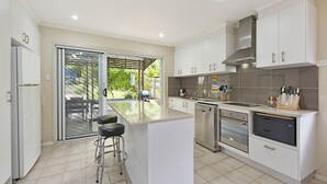 A modern kitchen with white cabinetry, stainless steel appliances, and a large island with bar stools. - 20 Roslin St Moffat Beach QLD (MOFFAT BEACH)