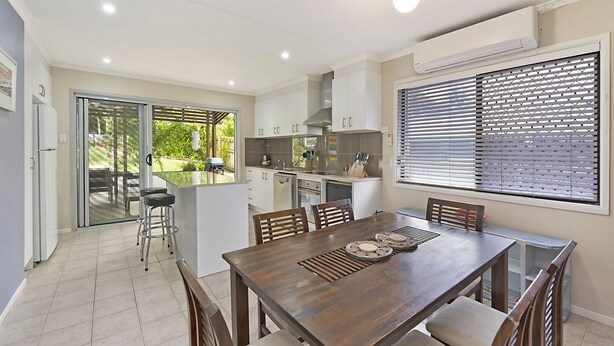 A modern kitchen with white cabinets, a dining area with a wooden table, and a sliding glass door leading to a patio. - 20 Roslin St Moffat Beach QLD (MOFFAT BEACH)