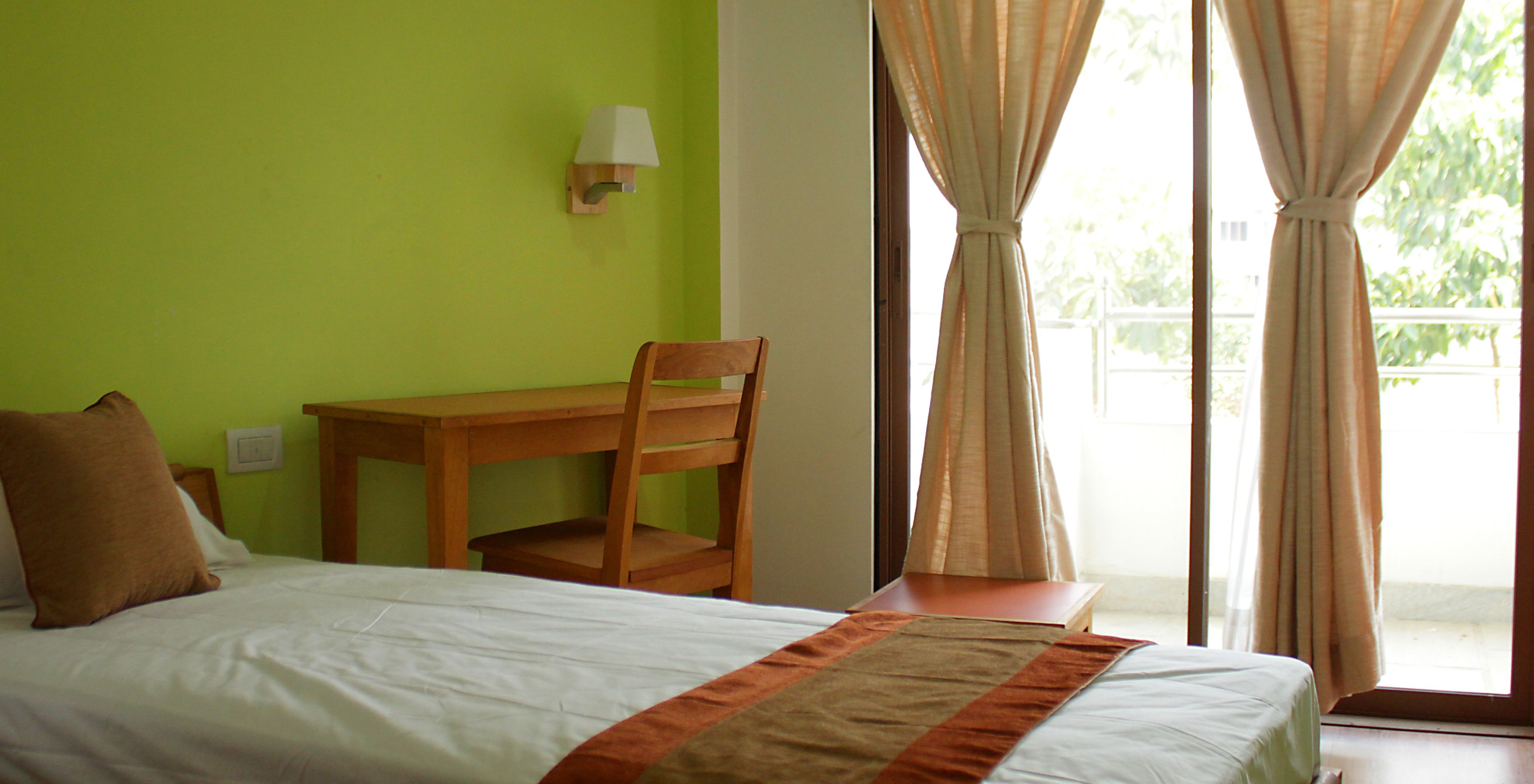 A room with a bed, a wooden desk, a chair, a lamp, and a view of the outdoors through a window with curtains.