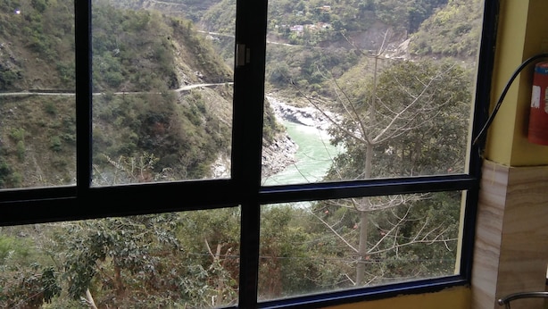 A room with a large window offering a view of a valley with a river. There are two red chairs and a table in the foreground. - Hotel Chakasha Govindam (Devaprayag)