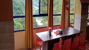 A room with a table and chairs, a window with curtains, and a view of a forested area. - Hotel Chakasha Govindam (Devaprayag)