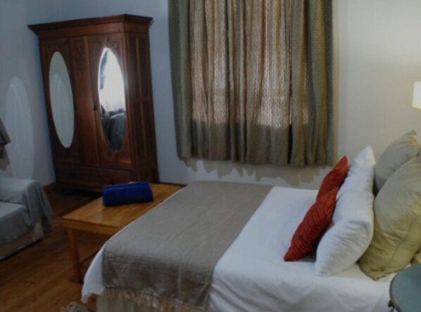 A bedroom with a bed, a wooden wardrobe, a small table, and a window with curtains.