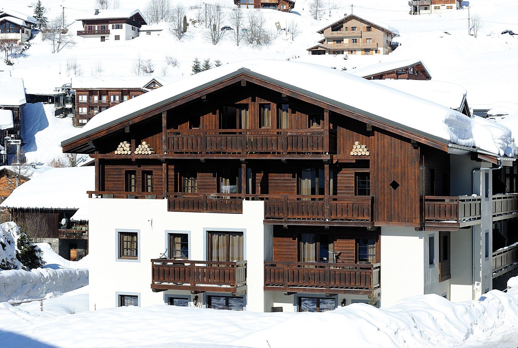 A multi-story wooden chalet with balconies, surrounded by snow-covered houses in a mountainous area.
