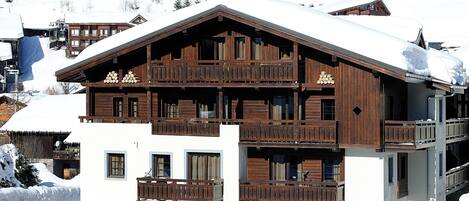 A multi-story wooden chalet with balconies, surrounded by snow-covered houses in a mountainous area.