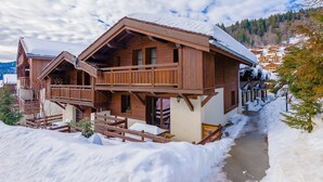 A wooden chalet with a balcony, surrounded by snow-covered ground and trees.