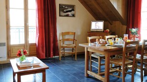 Un comedor con una mesa de madera dispuesta para el desayuno, un televisor sobre un soporte y una ventana con cortinas rojas.