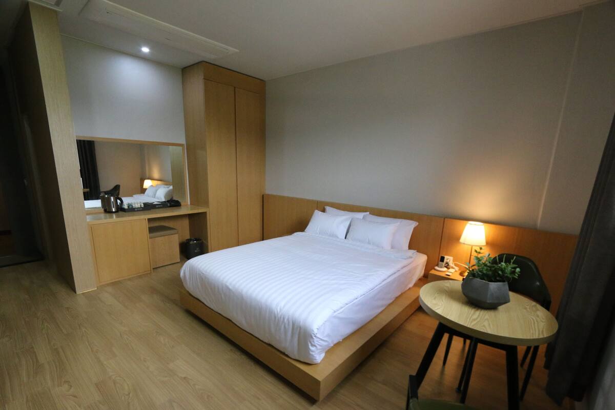 A hotel room with a large bed, a desk with a computer, a chair, and a small table with a plant.