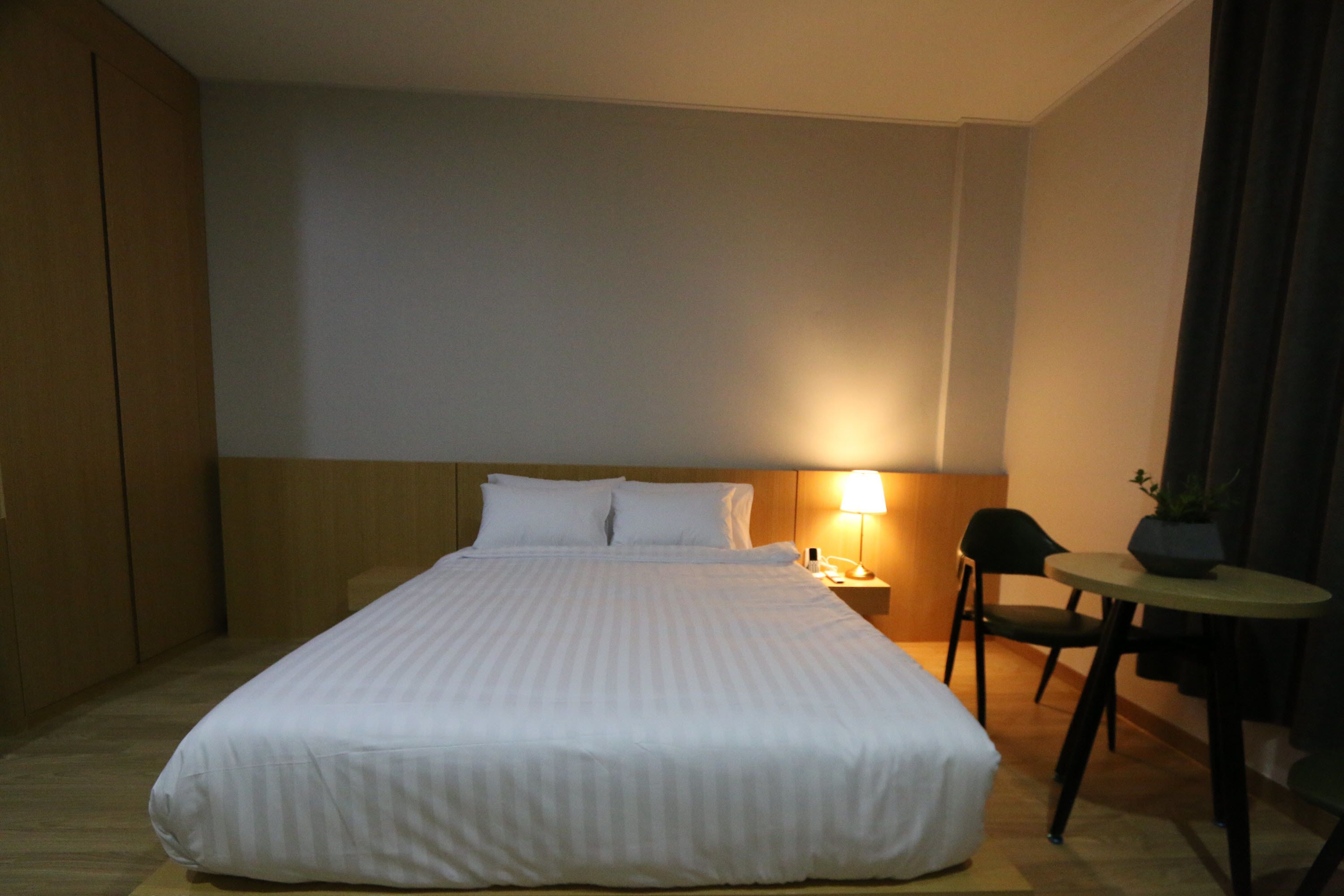 A hotel room with a large bed, a bedside table with a lamp, a chair, and a small table with a plant.