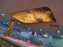 Un mural en el techo con figuras clásicas, una zona de recepción con mostrador y asientos, y un pilar con celosía metálica.
