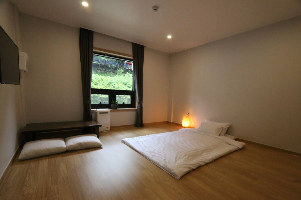 A modern bedroom with a large bed, a bedside table, a bench, and a window with a view of greenery.